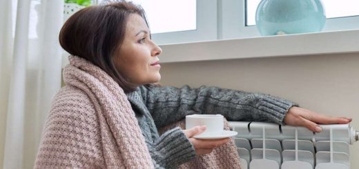 Kobieta o ciemnych włosach siedzi i ogrzewa się przy kaloryferze. Kobieta patrzy przez okno, przykryta jest beżowym kocem i trzyma rękę na kaloryferze, w drugiej ręce trzyma filiżankę z herbatą.