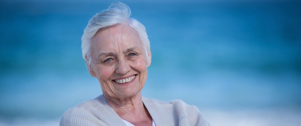 Uśmiechnięta starsza kobieta siedzi na plaży, w tle widać morze