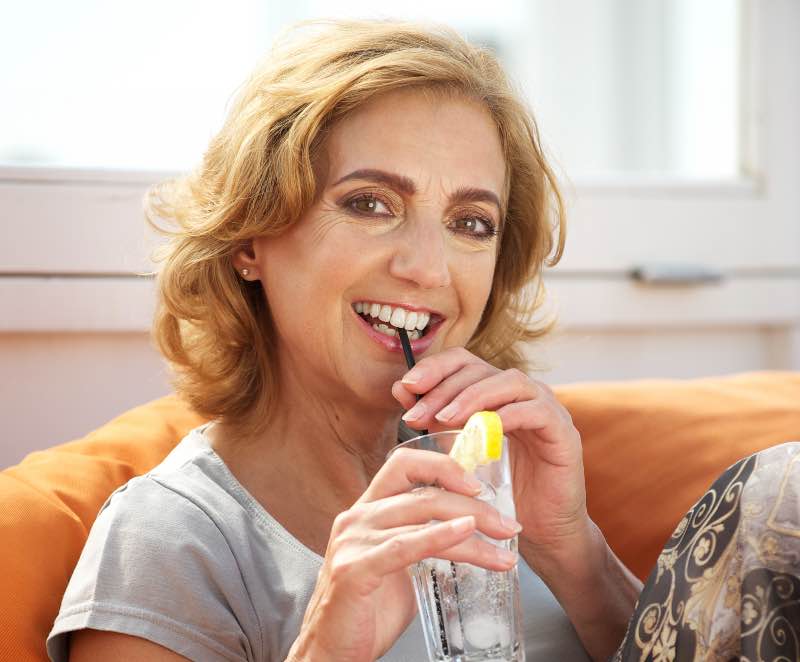Dojrzała kobieta siedzi na kanapie i pije wodę z cytryną przez rurkę