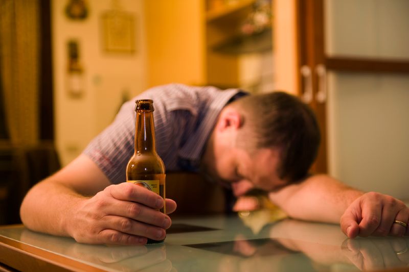 Mężczyzna z piwem w dłoni śpi z głową na stole.