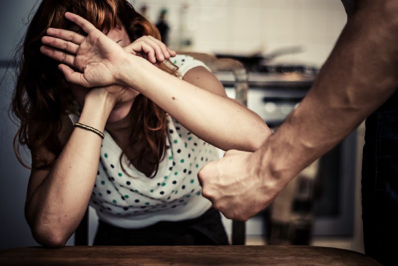 Kobieta siedząca przy stole i broniąca się rękoma przed pobiciem przez mężczyznę, który stoi nad nią z zaciśniętą pięścią.
