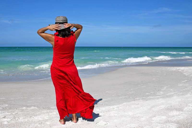 Dojrzała kobieta w czerwonej sukni i słomkowym kapeluszu stoi na słonecznej plaży tyłem do obiektywu