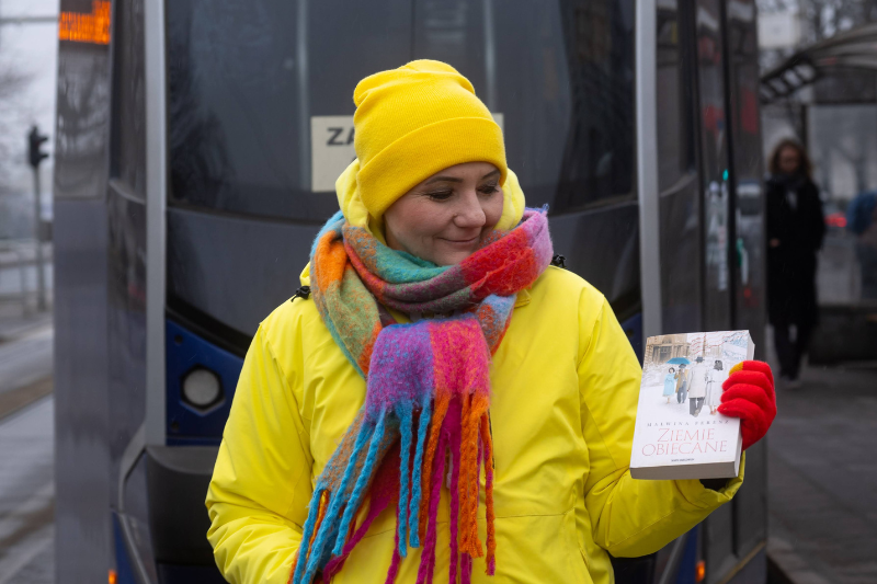 Malwina Fereny na tle tramwaju, w rękach trzyma swoja książkę "Ziemie obiecane"
